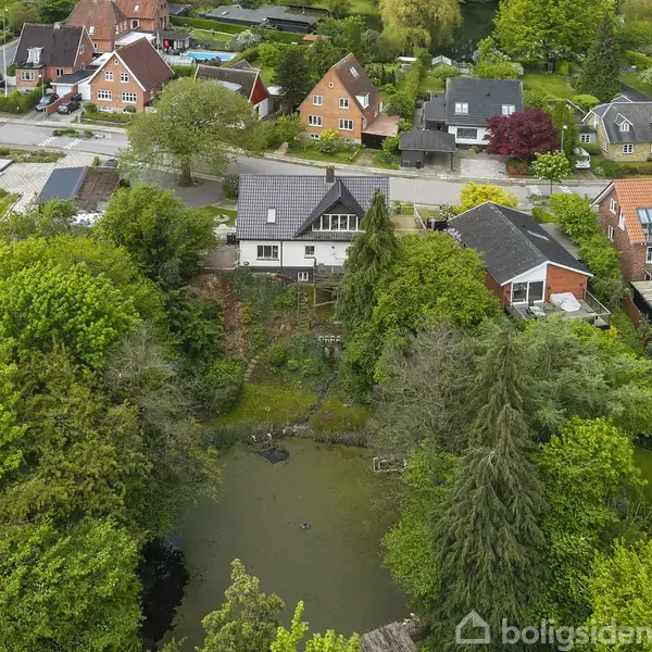 Boliger omgivet af frodig vegetation og en dam. De røde og hvide huse ligger langs en vej i et grønt boligområde med store træer og haver.