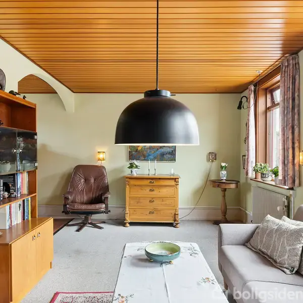 Sort loftslampe hænger over sofabord i stue med grå sofa, trævægpanel, lænestol og vitrineskab. Vindue med blomster og gardiner, kommode med maleri over. Hyggelig indretning.