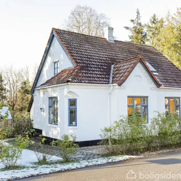 Et hvidt hus med brunt tegltag står stille om vinteren, omgivet af snebeklædt have og høje træer. Husets vinduer reflekterer sollys.