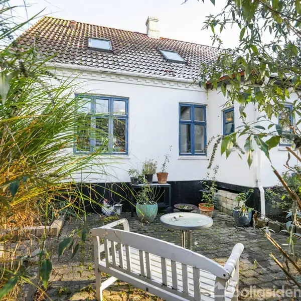 Et hvidt hus med blå vinduer står i en haveomgivelse. Foran huset er der en terrasse med en hvid bænk, runde havebord og potteplanter. Træer og buske omgiver området.