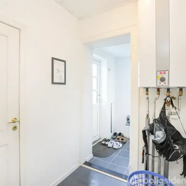 En varmepumpe hænger på væggen, omgivende af en hvidmalet gang. Til højre ses et par tasker og en blå kurv, og i baggrunden står flere par sko ved en dør.