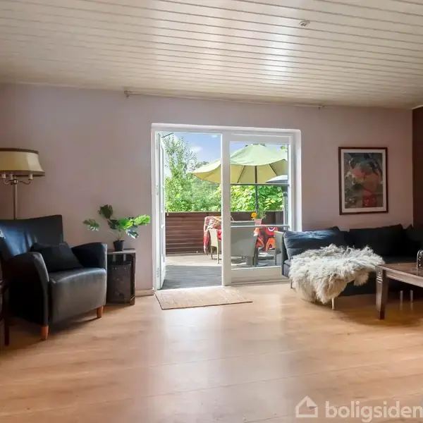 Stue med sort læderstol og sofa dækket af fåreskind. Store glasdøre fører ud til terrasse med parasol og stole, omgivet af grønt. Vægge har kunst og hylder.
