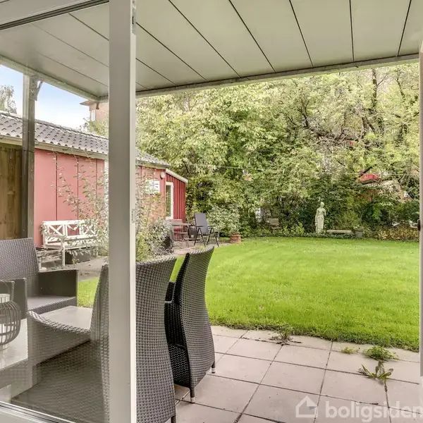 En glasdør står åben og afslører en terrasse med flettede møbler, der står på fliser. Græsplænen strækker sig mod en rød træbygning og en frodig, grøn have med træer.