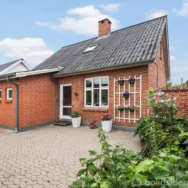 Rødt murstenshus med gråt tegltag står i en hyggelig gårdhave. Flere blomsterpotter og grøn vegetation omringer området, med en stengang, i et beboelsesområde under en blå himmel.