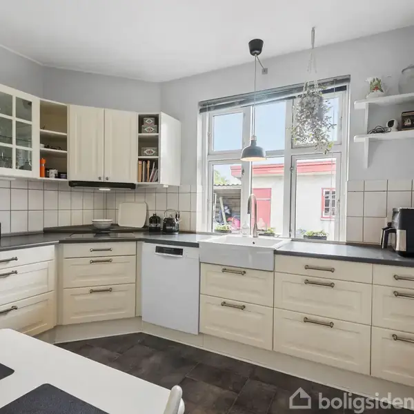 Et køkken med lyse træskabe og sorte bordplader. Der står en opvaskemaskine og en kaffemaskine. Et vindue med lamper hænger over vasken, og udenfor ses et rødt hus.