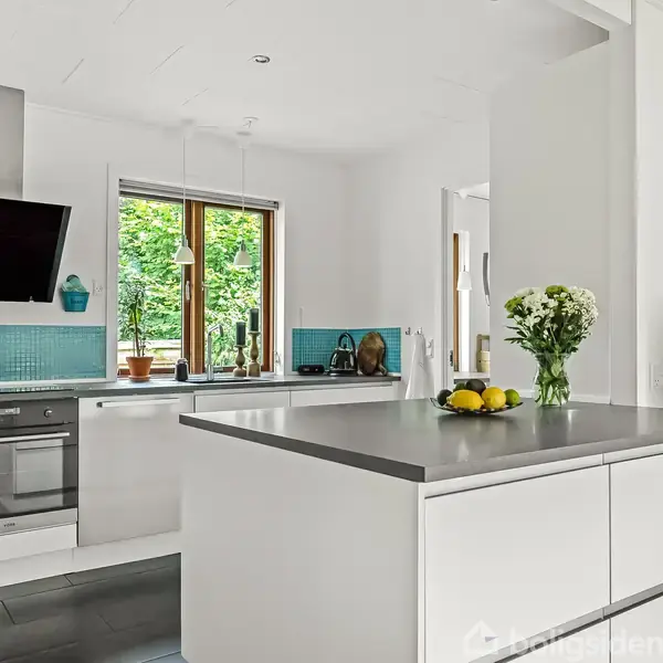 Køkken med moderne hvidt interiør, der har en grå bordplade. Blomster og citroner er placeret på øen. Rummet har store vinduer med udsigt til grønne træer.