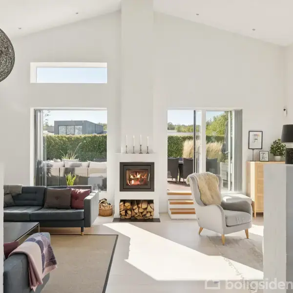 En moderne stue med en mørk sofa og lænestol, hvor en pejs brænder lystigt. Store vinduer lukker lys ind, og en dør fører ud til terrassen. En farverig kunstplakat hænger på væggen.