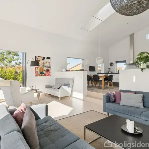 Stue med moderne møbler, to grå sofaer og et spisebord i baggrunden. Store vinduer slipper naturligt lys ind. Planter og kunstværker dekorerer rummet, der har en lys og åben atmosfære.
