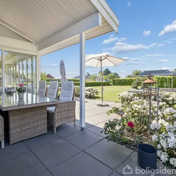 En overdækket terrasse med en stor glasbord og fletstole står ved siden af en velplejet have med blomster og en parasol. Huset har store vinduespartier og blå himmel ses.
