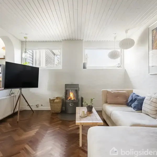 Stue med moderne sofa foran et sofabord. Et vægmonteret TV hænger til venstre, og en brændeovn står ved vinduet. Lyse vægge og trægulv skaber en hyggelig atmosfære.