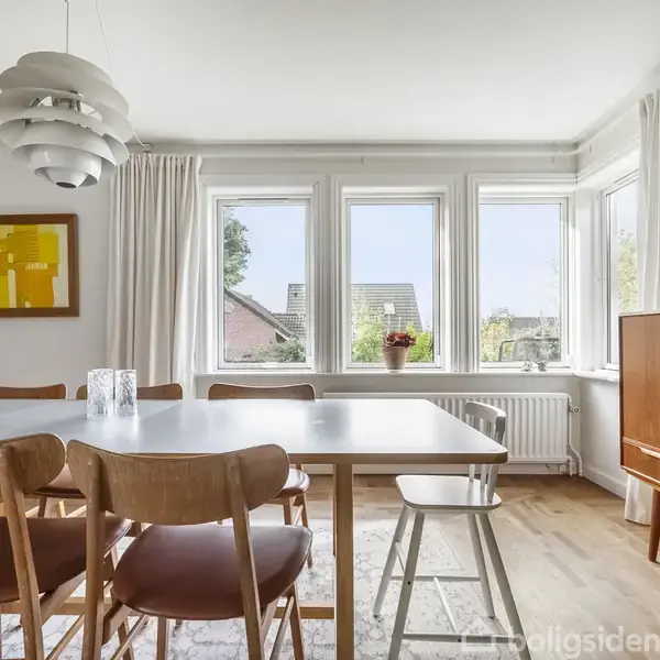 Et spisebord med seks træstole står i et lyst rum. På væggen hænger et farverigt billede. Store vinduer i baggrunden giver udsigt til et hus og grønt landskab.