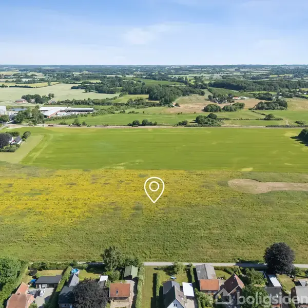 En stor, grøn mark strækker sig i et åbent landskab med spredt vegetation. Foran markerne er der nogle huse. En hvid markør angiver et sted midt på marken.