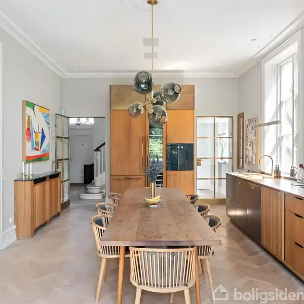 Et spisebord med moderne stole står i et lyst køkken med træskabe og en grøn marmorvask. Kunstværker hænger på væggene, og en unik loftslampe hænger over bordet.