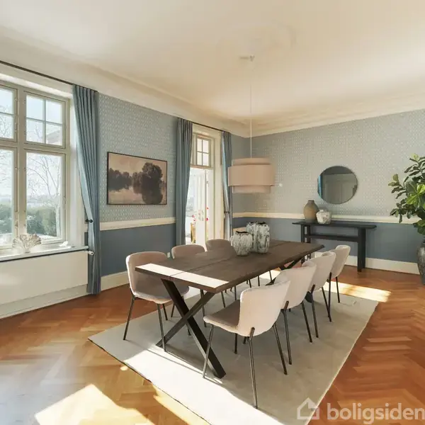 Spisebord med otte stole står i et lyst rum med blå vægge og træparketgulv. Store vinduer og døre lukker naturligt lys ind. En stor plante og spejl pryder rummet.