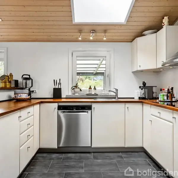 Lyst køkken med hvide skabe, brun bordplade og en indbygget vask. Der er en opvaskemaskine og et loftsvindue. Udenfor ses træer gennem vinduerne. Flere køkkenredskaber og en ovn er synlige.