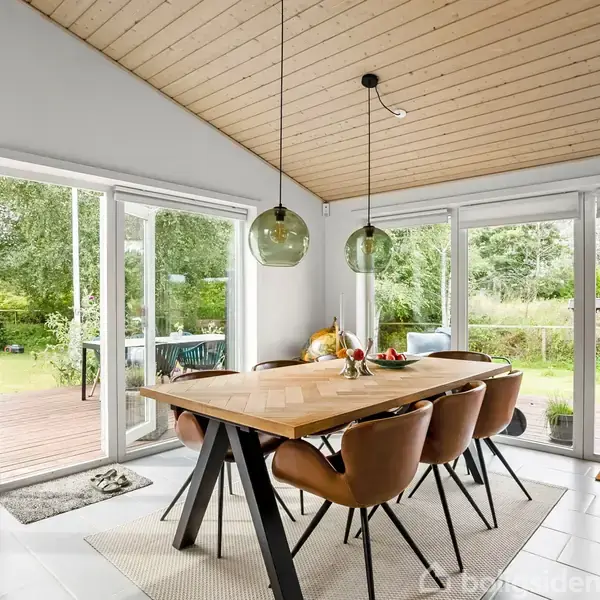 Lang spisebord omgivet af brune stole står i en lys spisestue med store vinduer, der giver udsigt til en frodig have. Teksten "HYGGE" ses på en dørmåtte.