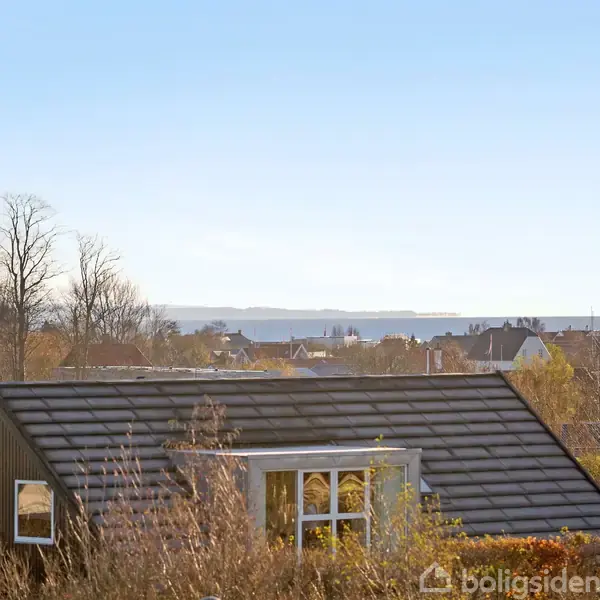 Et hus med mørkt tag ligger omgivet af træer, med udsigt over en by og havet i horisonten under en klar himmel.