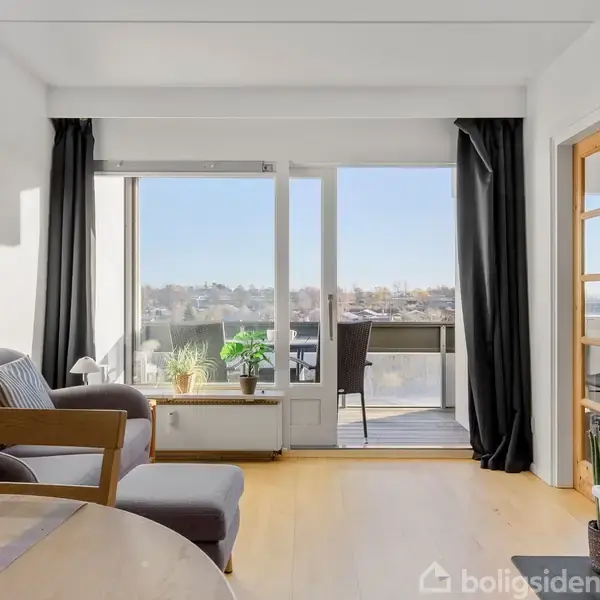 En stue med en grå sofa og et bord står ved siden af en stor glasdør. Udenfor er der en balkon med stole, planter og udsigt over byen.