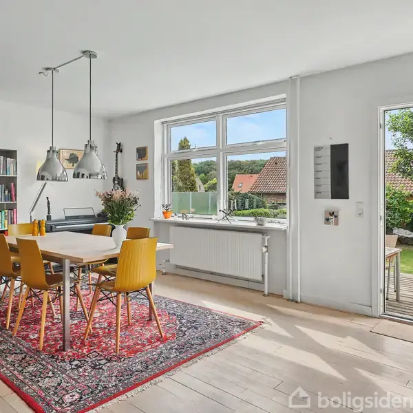 Et moderne spisebord med gule stole står på et rødt orientalsk tæppe. Ved siden af er en bogreol fyldt med bøger. Rummet åbner ud til en have gennem en dør.