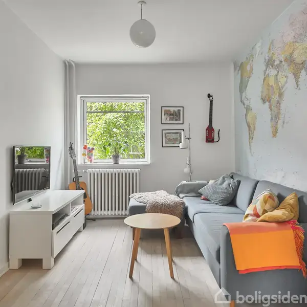 En stue med en grå sofa fyldt med puder, placeret mod en væg med et verdenskort. Et lille rundt bord foran sofaen. Et vindue med grøn udsigt og en tv-bænk står modsat sofaen.