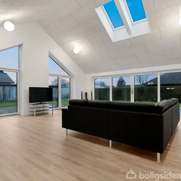 Sort sofa står i en lys stue med trægulv og hvide vægge. Store vinduer og et opdelt loft med ovenlysvinduer giver udsigt til en have og blå himmel.