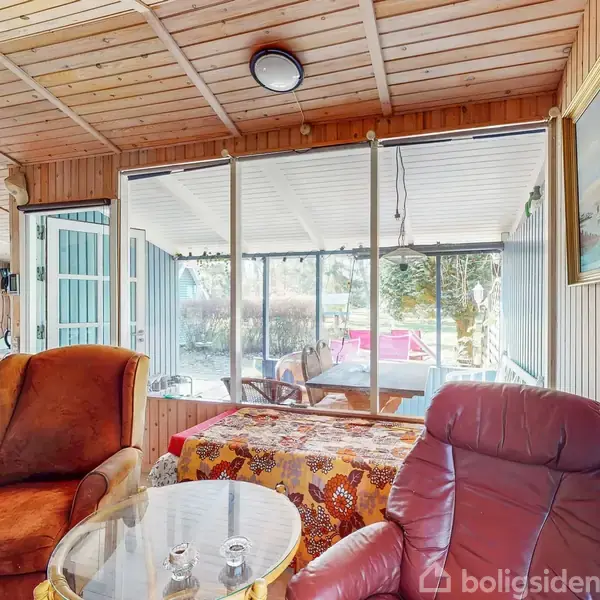 To lænestole står ved et lille rundt bord i et rum med træbeklædning. Store vinduer viser en terrasse udenfor. Et maleri hænger på væggen.