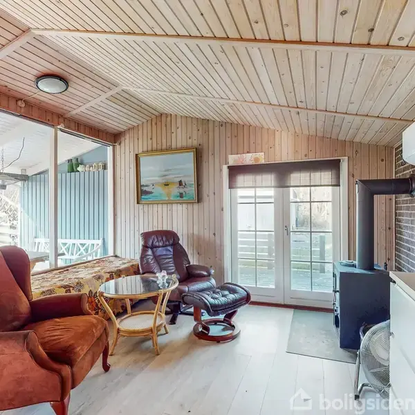 En stue indrettet med en brun lænestol og gyngestol ved et rundt bord. En pejs står til højre, og store vinduer åbner mod en terrasse. Trævægge og loft skaber en hyggelig atmosfære.