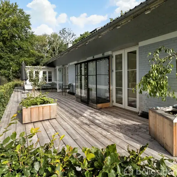 Terrasse med trædæk omkranset af planter; store vinduer fører ind til et hus. Planter i træbeholdere står langs kanten, og haven ses med grønt græs og træer i baggrunden.