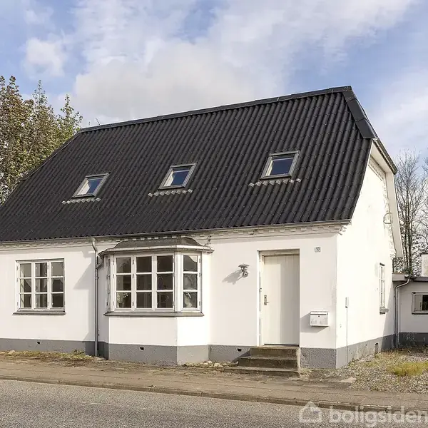 Hvidt hus med sort tag og to etager står stille på en stille villavej. Forhaven har en lille trappesten ved indgangen, og omgivelserne har nogle træer og en vejlampe.