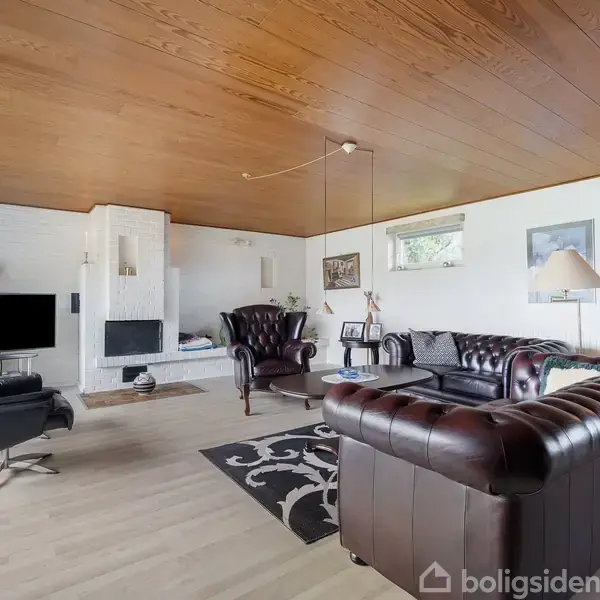 Brune lædersofaer er opstillet omkring et tæppe i en stue med trægulv og loft. I hjørnet er en pejs, flankeret af en sort lænestol og et tv.