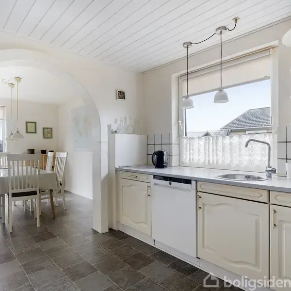 Et lyst køkken med hvide skabe, der åbner mod en spisestue gennem en buegang. To pendellamper hænger over vasken nær et stort vindue, der giver udsigt til et hus.