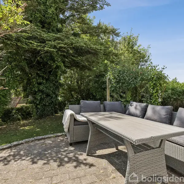 Gråt havemøbelsæt står på en flisebelagt terrasse. Møblerne består af en sofa, stole og et bord. Omkring er der grønne træer og buske under en blå himmel.