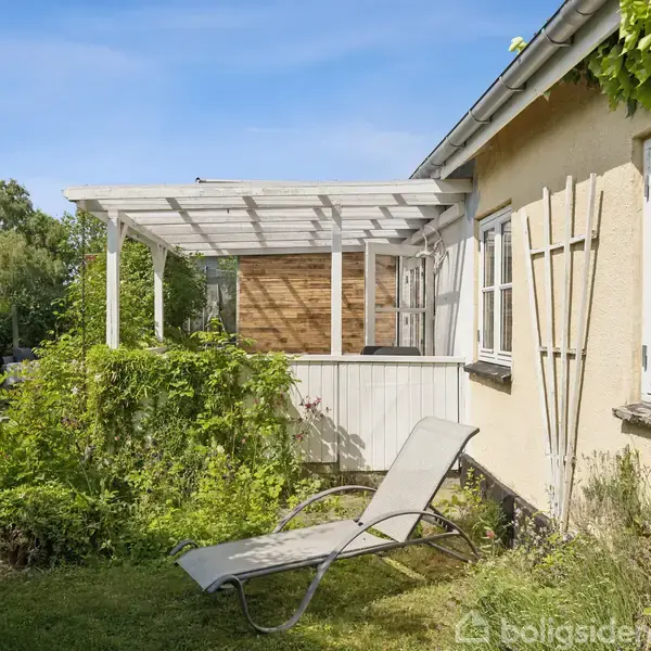 En liggestol står ved en solrig terrasse af træ, omkranset af grønne buske og græsplæne, tilknyttet et hus med lyse ydervægge og hvide vinduer.