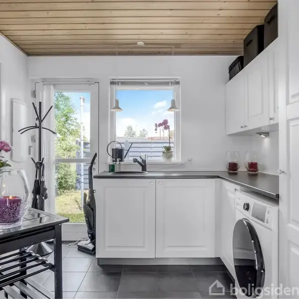 Køkken har hvide skabe og en sort bordplade. Maskiner står ved siden af vindue. Lysindfald fra terrassedør afslører en grøn have udenfor. Ingen tekst fundet.