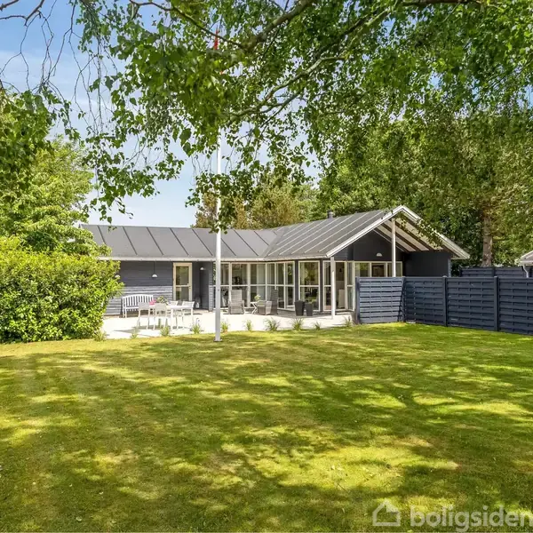 Et moderne, mørkt træhus med store vinduer står i en velplejet have. En terrasse med havemøbler flankerer huset, og høje træer omkranser grunden under en blå himmel.