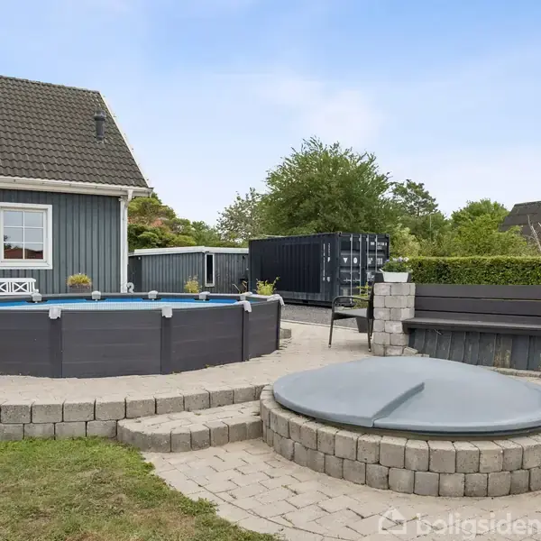 Et rundt, hævet svømmebassin står i en have foran et gråt træhus. Der er en flisebelagt sti, græsplæne og en overdækket jacuzzi i nærheden.