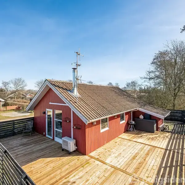 Rødt hus med skråt tag står på en stor træterrasse, omgivet af et hegn og grønne træer. Klar blå himmel vises i baggrunden.