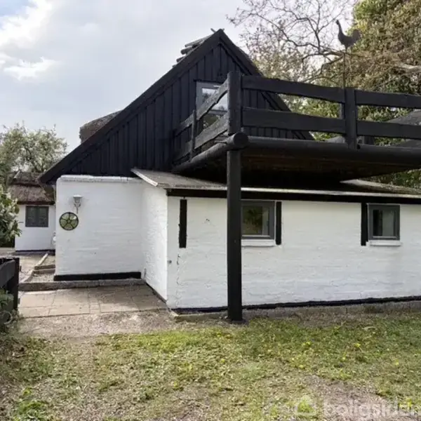 Et hvidt hus med mørkt tag har en stor træterrasse ovenpå. Det står i en grøn have med træer og flisebelagt område ved siden af.