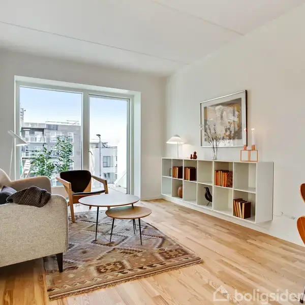 Stue med en grå sofa til venstre, en reol med bøger og pyntegenstande i midten, og et bord med stole til højre. Store vinduer giver udsigt til bygninger udenfor.