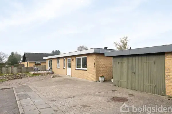 Et gult murstenshus med fladt tag har grøn garageport. Huset står i en åben forstadshave med asfalteret indkørsel og spredt beplantning. I baggrunden ses nabohus og træer.