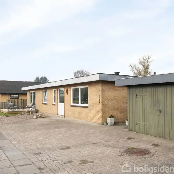 En gulstensvilla med fladt tag står på en flisebelagt indkørsel. Til højre ses en grøn garageport. Omgivelserne inkluderer en have og et nabohus. Nummeret 51 ses ved døren.
