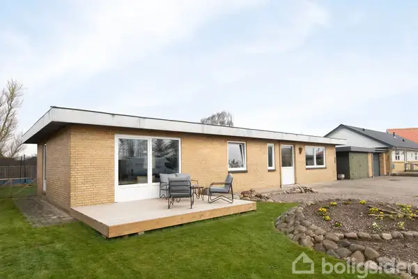 Et murstenshus med fladt tag står stille i en have med græsplæne og blomsterbed. Terrassen foran huset har et bord og stole.