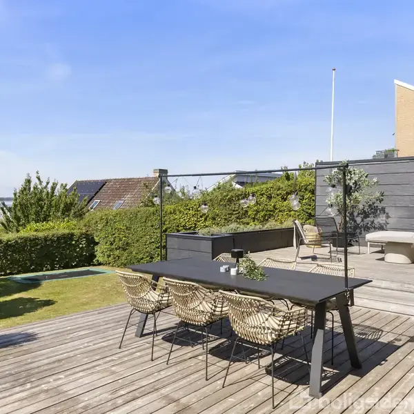 En sort spisebord med fire stole står på en træterrasse. En lav planter er ved siden af bordet. En sofa står i baggrunden ved et hegn, omgivet af grønne buske.
