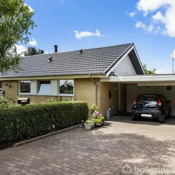 Et hus med skråtag står ved siden af en indkørsel. En sort bil holder parkeret under en carport. Haven er velplejet med hække, grusbelægning og blomsterkrukker.