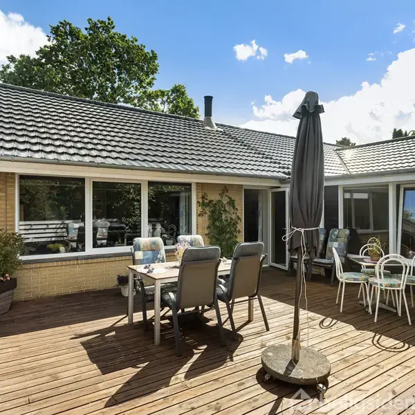 Terrasse med trædæk indeholder spisebord og stole samt en parasol. Baggrunden viser en gul murstenshusfacade med store vinduer. Træer og blå himmel er synlige i baggrunden.