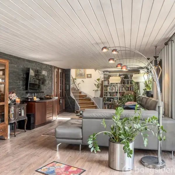 En stue med en grå hjørnesofa og en moderne gulvlampe. Et stort vindue giver udsigt til en have. En trappe fører op til en bogreol ved siden af en skab.