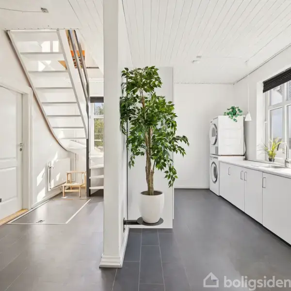 Stueetage med hvid trappe til venstre, plante i midten, og køkken på højre side med vaskemaskine, tørretumbler og hvide skabe. Lyse vægge og sorte fliser skaber en moderne atmosfære.