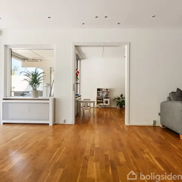 Stue med parketgulv, hvor en grå sofa står til højre. Stuen har store vinduer, der leder ud til en terrasse. En indendørs plante står nær et spejl til venstre.