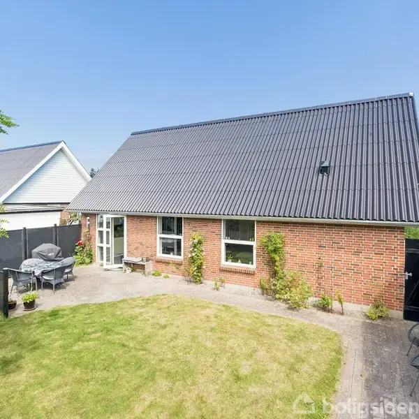 Rødt murstenshus med sort tag står stille i sollys. Forhave med græsplæne og terrasse til højre, omkranset af hegn. Blåt skyfrit himmelloft giver atmosfære.