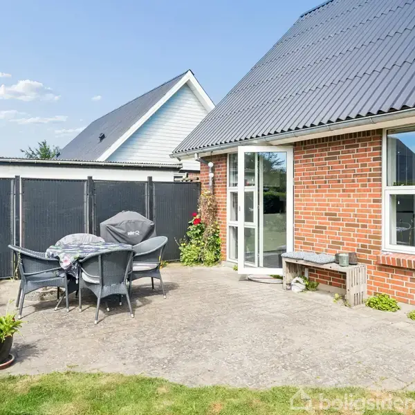 En udendørs terrasse med et bord og seks stole dækket af betræk. Omgivet af en rød murstensvæg og hegn. Blomsterkrukker og græsplæne i baggrunden.